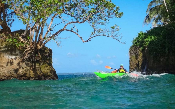 A person paddles a kayak through blue water between two tall rocks covered with trees and greenery.